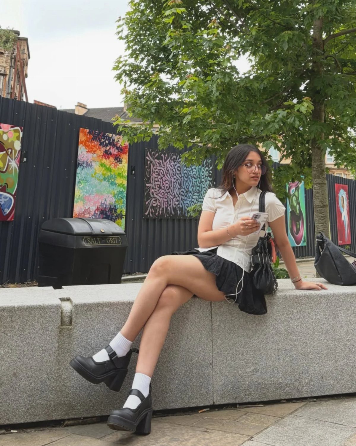 A young woman wearing a white shirt, black pleated skirt, and platform shoes sitting on a ledge.